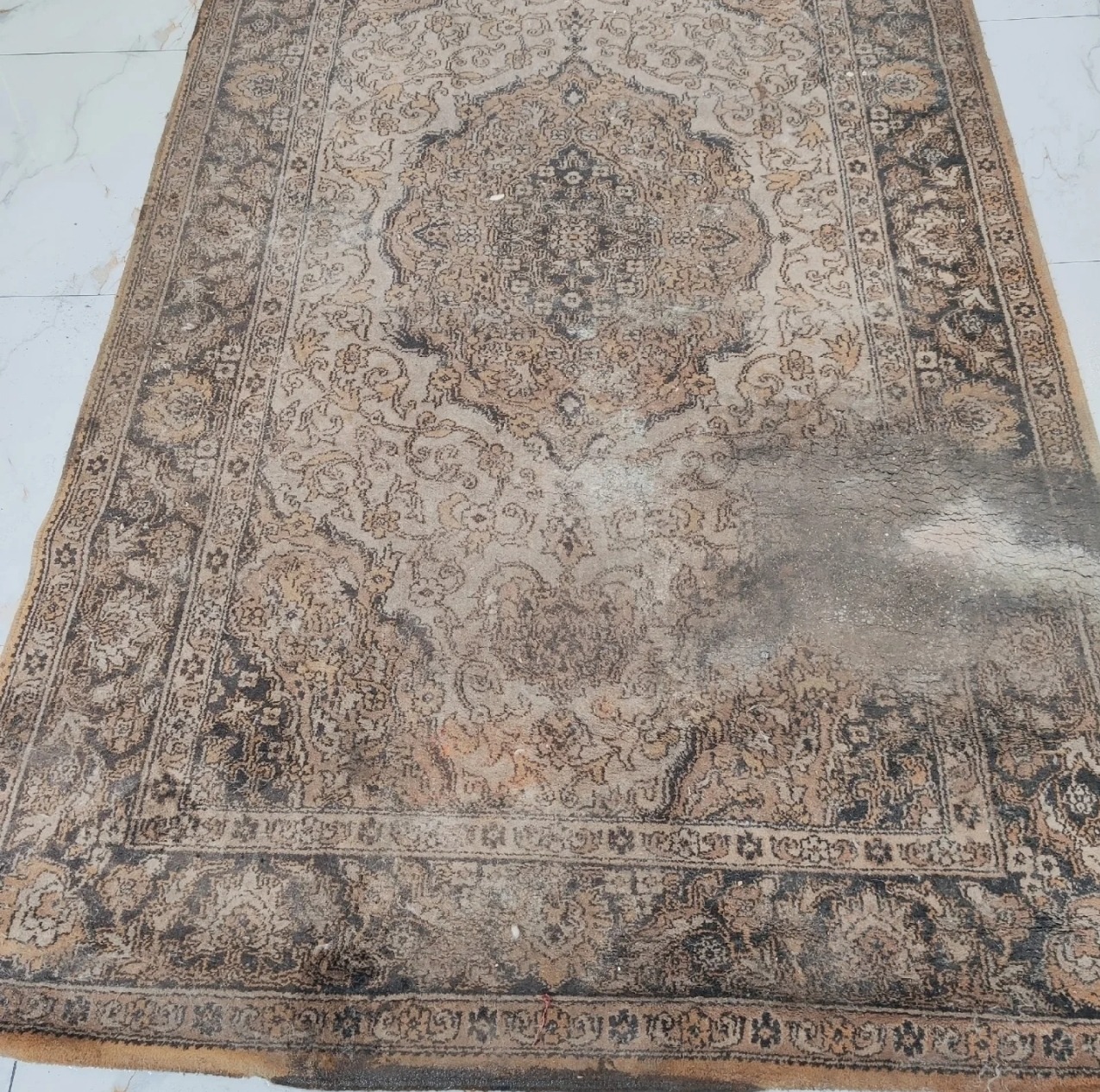 Zabrudzony, beżowo-brązowy dywan orientalny z wyraźnymi plamami i śladami użytkowania, leżący na jasnej podłodze. Widoczne detale wzoru i uszkodzenia.