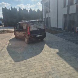 Golden-Bruk Rafał Czyż - Ułożona kostka brukowa na podjeździe z zaparkowanym autem firmy brukarskiej. Widoczny budynek w tle i fragment przygotowanego terenu pod dalsze prace.