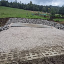 Golden-Bruk Rafał Czyż - Przygotowany teren pod budowę z wysypanym żwirem i obłożony kamieniami, widok na zielone wzgórze i las w tle, pochmurne niebo.