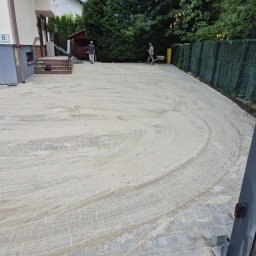 Golden-Bruk Rafał Czyż - Układanie kostki brukowej: wyrównany teren piaskiem kwarcowym, przygotowany pod ułożenie kostki. Widoczni pracownicy i budynek w tle.