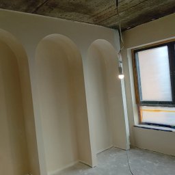 Dima Potapow - Wnętrze po remoncie: kremowe ściany z trzema wnękami w kształcie łuków, okno zabezpieczone folią malarską, surowy betonowy sufit i wisząca żarówka.