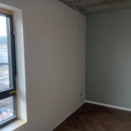 Dima Potapow - Świeżo pomalowane wnętrze z parkietem w jodełkę. Ściany w odcieniach szarości i zieleni, widoczne gniazdka elektryczne i okno z taśmą malarską.