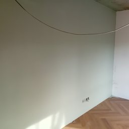 Dima Potapow - Świeżo pomalowane wnętrze z parkietem w jodełkę. Widoczne gniazdka elektryczne i surowy betonowy sufit. Kabel zwisa z góry, a żarówka oświetla róg pomieszczenia.