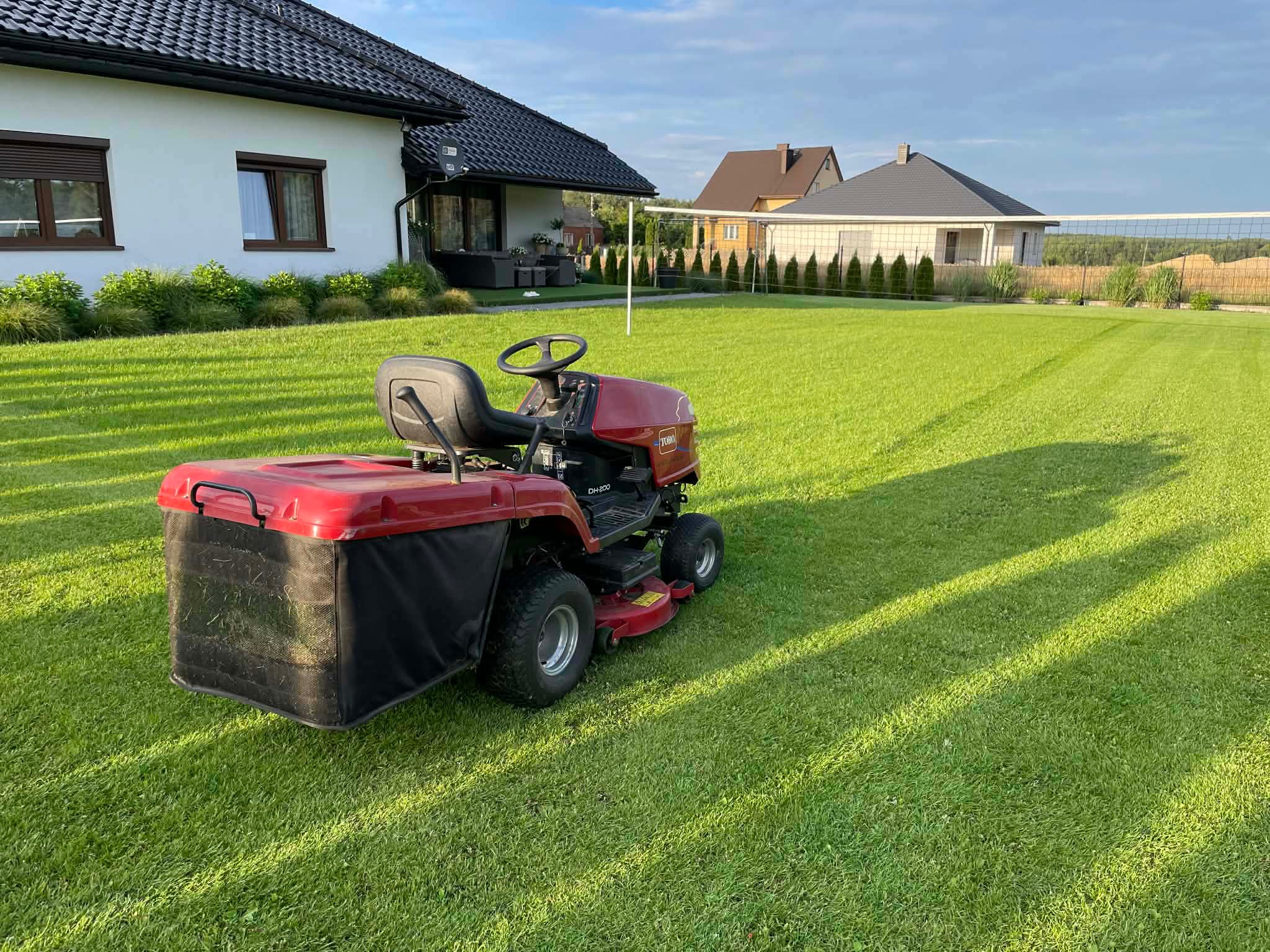 Czerwona kosiarka traktorek na tle idealnie przystrzyżonego trawnika przed domem z siatką do siatkówki. Perfekcyjny trawnik.