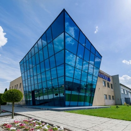 Nowoczesny budynek biurowy z fasadą ze szkła refleksyjnego, błękitne niebo odbija się w oknach. Widok z dołu, zieleń i kwiaty na pierwszym planie.