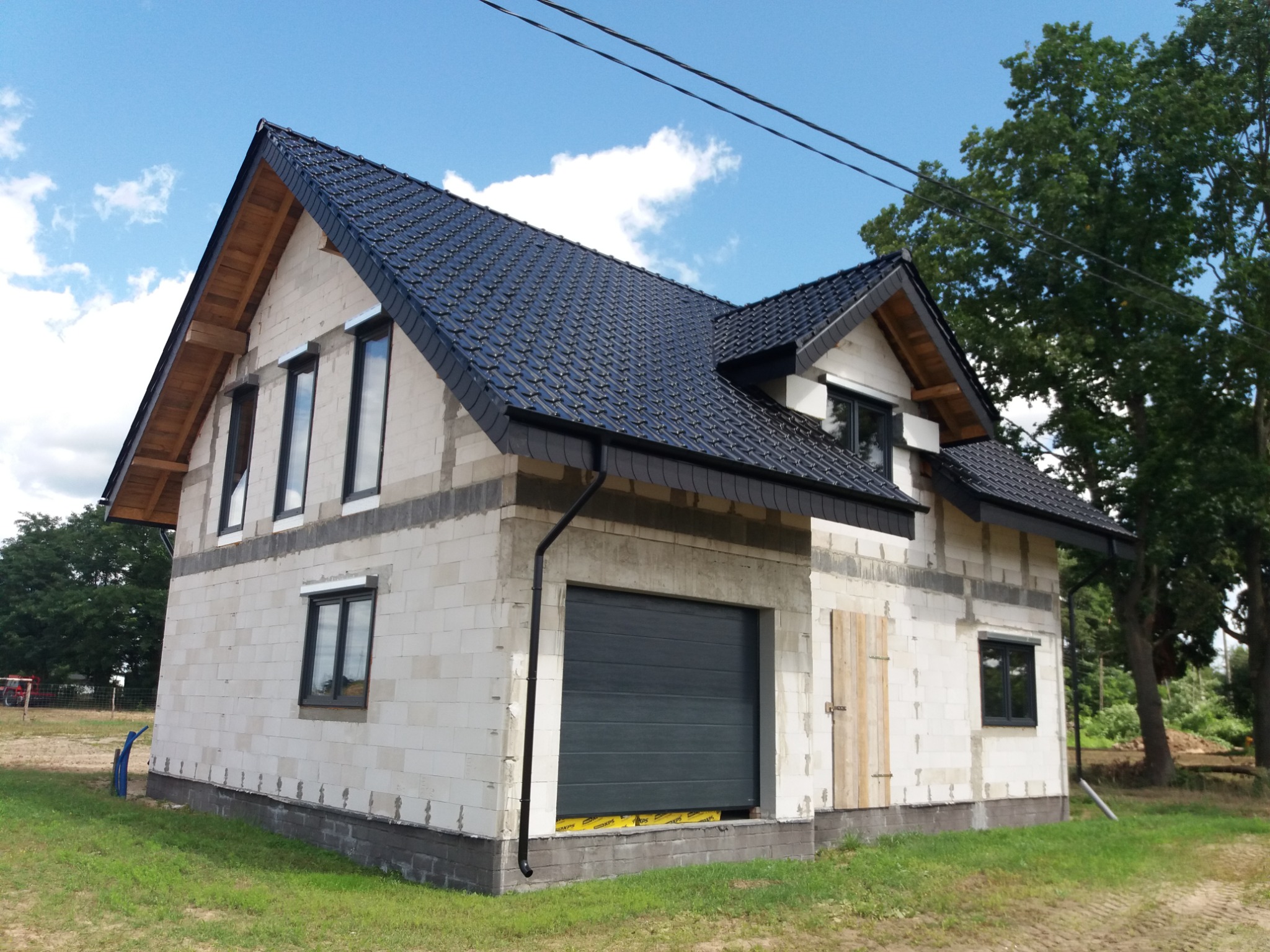 Surowy dom jednorodzinny w trakcie budowy: czarne okna, brama garażowa i dachówka kontrastują z białymi bloczkami. Widoczne elementy konstrukcji dachu.
