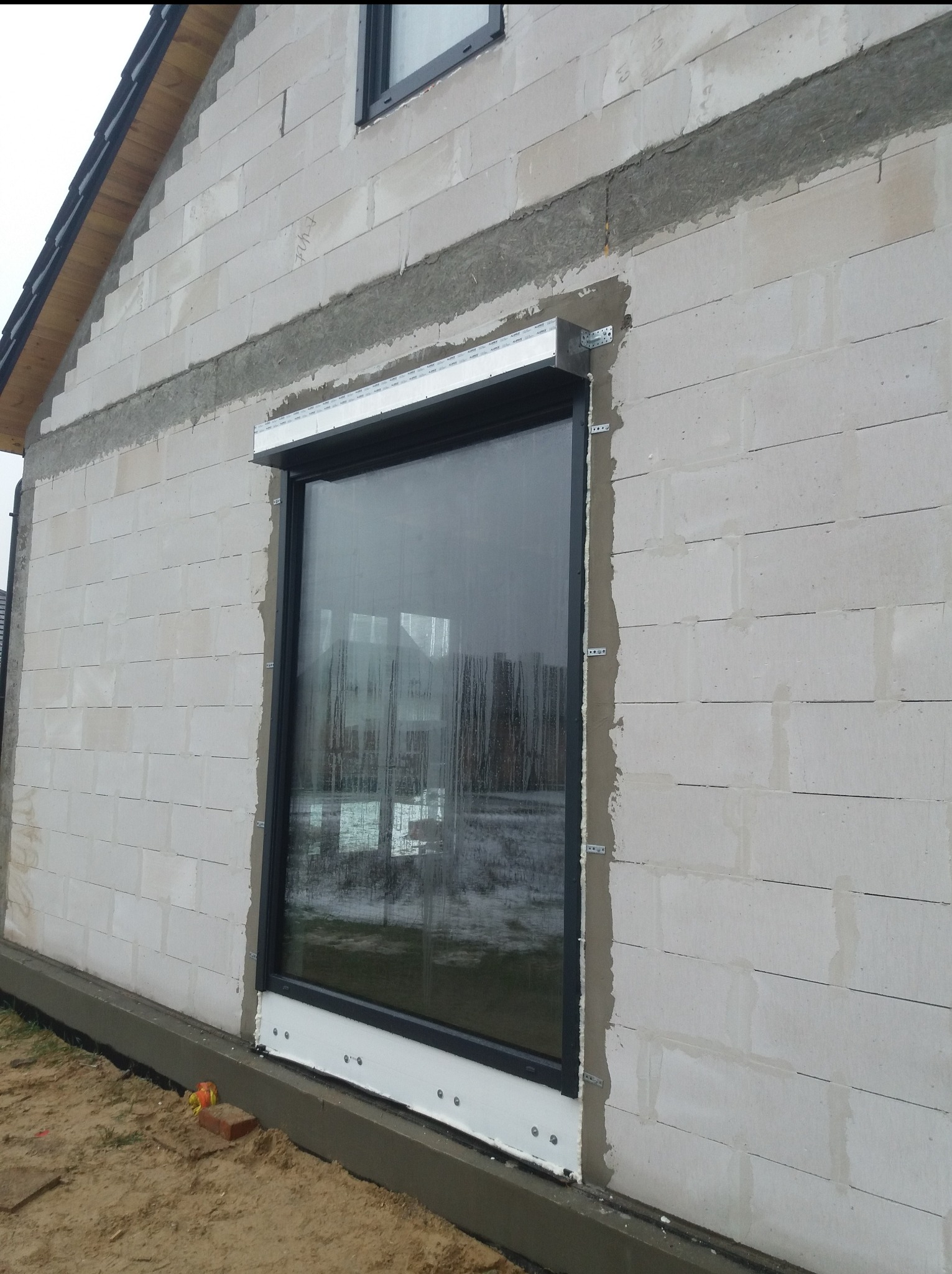 Montaż okna w surowym murze z betonu komórkowego. Widoczne metalowe mocowania i pianka montażowa. Okno z czarną ramą, jeszcze bez wykończenia.