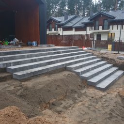 SIMPLA Marcin Skorulski - Schody z szarych, betonowych bloków prowadzące do nowoczesnego domu. W tle widoczny las i częściowo ogrodzenie. Na pierwszym planie ziemia i mała dziewczynka.