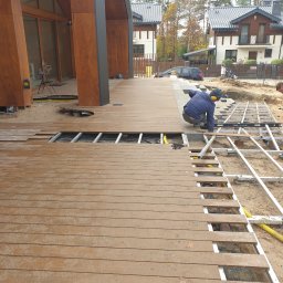 SIMPLA Marcin Skorulski - Montaż drewnianego tarasu przy domu jednorodzinnym. Widoczne deski tarasowe, konstrukcja nośna i pracownicy podczas układania. Częściowo ułożona podłoga tarasu.