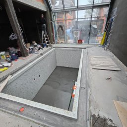 SIMPLA Marcin Skorulski - Wnętrze basenu w trakcie budowy z mozaiką szkliwioną, widoczne narzędzia i pracownik na drabinie. Duże okno z widokiem na zewnątrz. Etap doszczelniania przepustów w żelbecie.