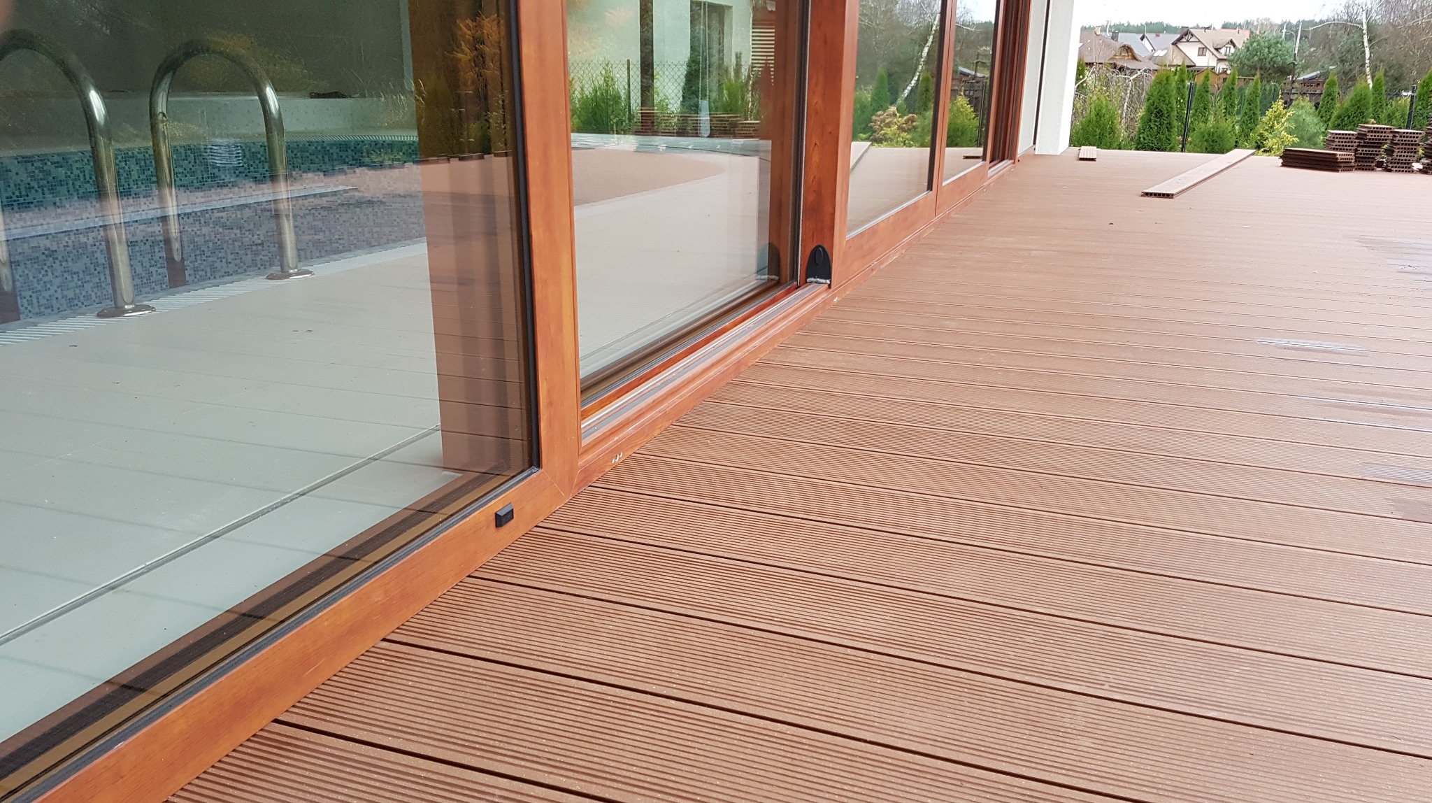 Kompozytowy taras w odcieniach brązu, przylegający do budynku z dużymi oknami, za którymi widoczny jest basen i elementy krajobrazu.