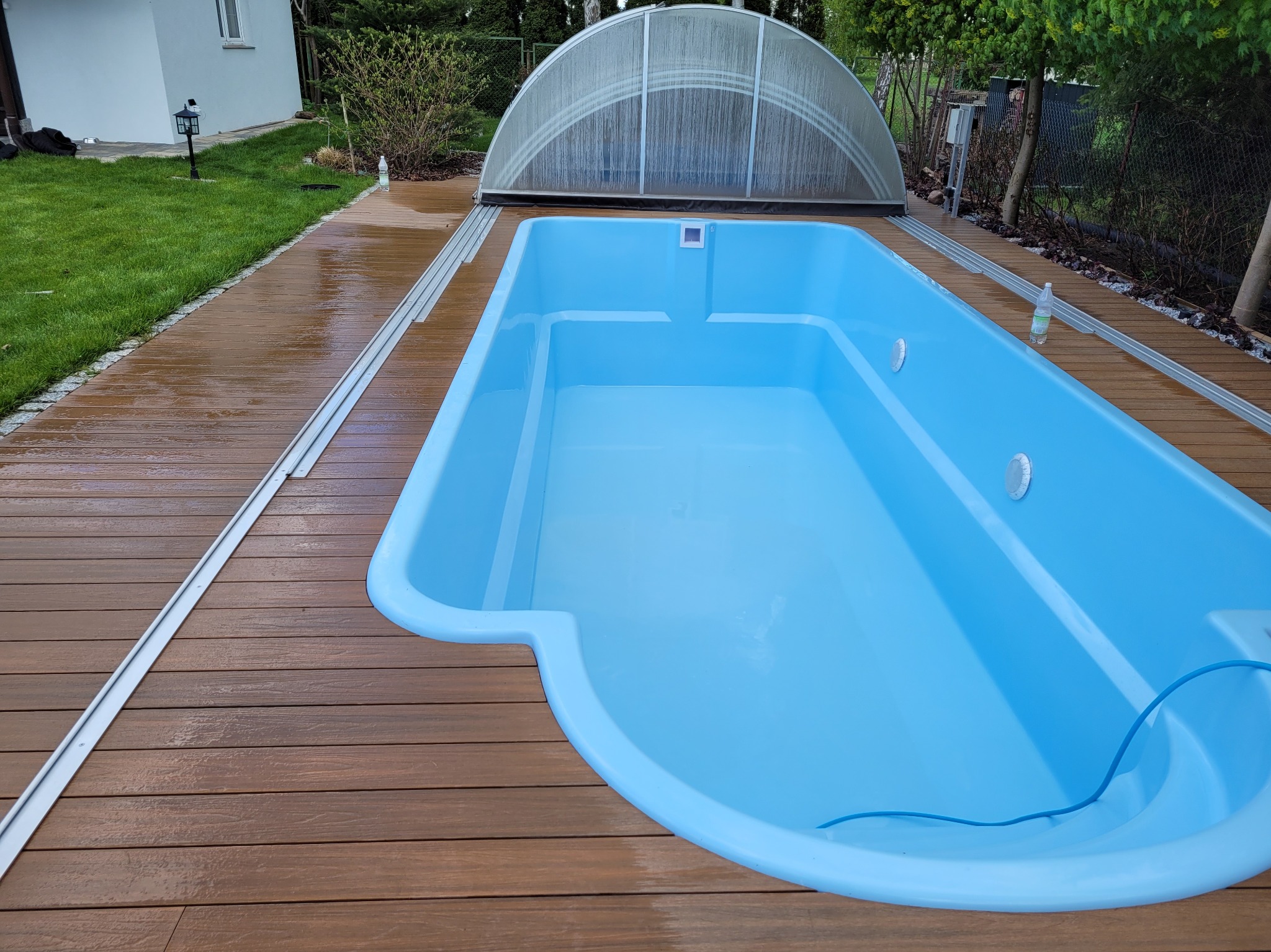 Niebieski basen z zadaszeniem na drewnianym tarasie, otoczony zielenią. Widoczne detale wykończenia i instalacji basenowej. Ujęcie z góry.