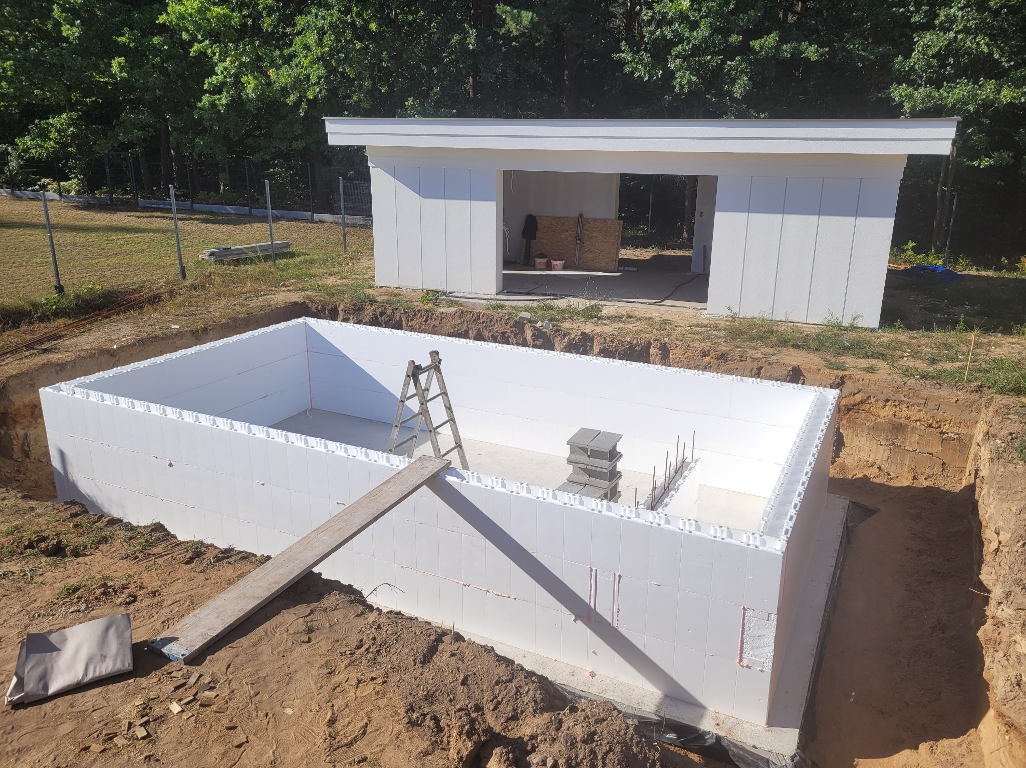 Budowa basenu: białe ściany z izolacji, drabina w środku wykopu, w tle budynek gospodarczy. Widok na plac budowy w słoneczny dzień.