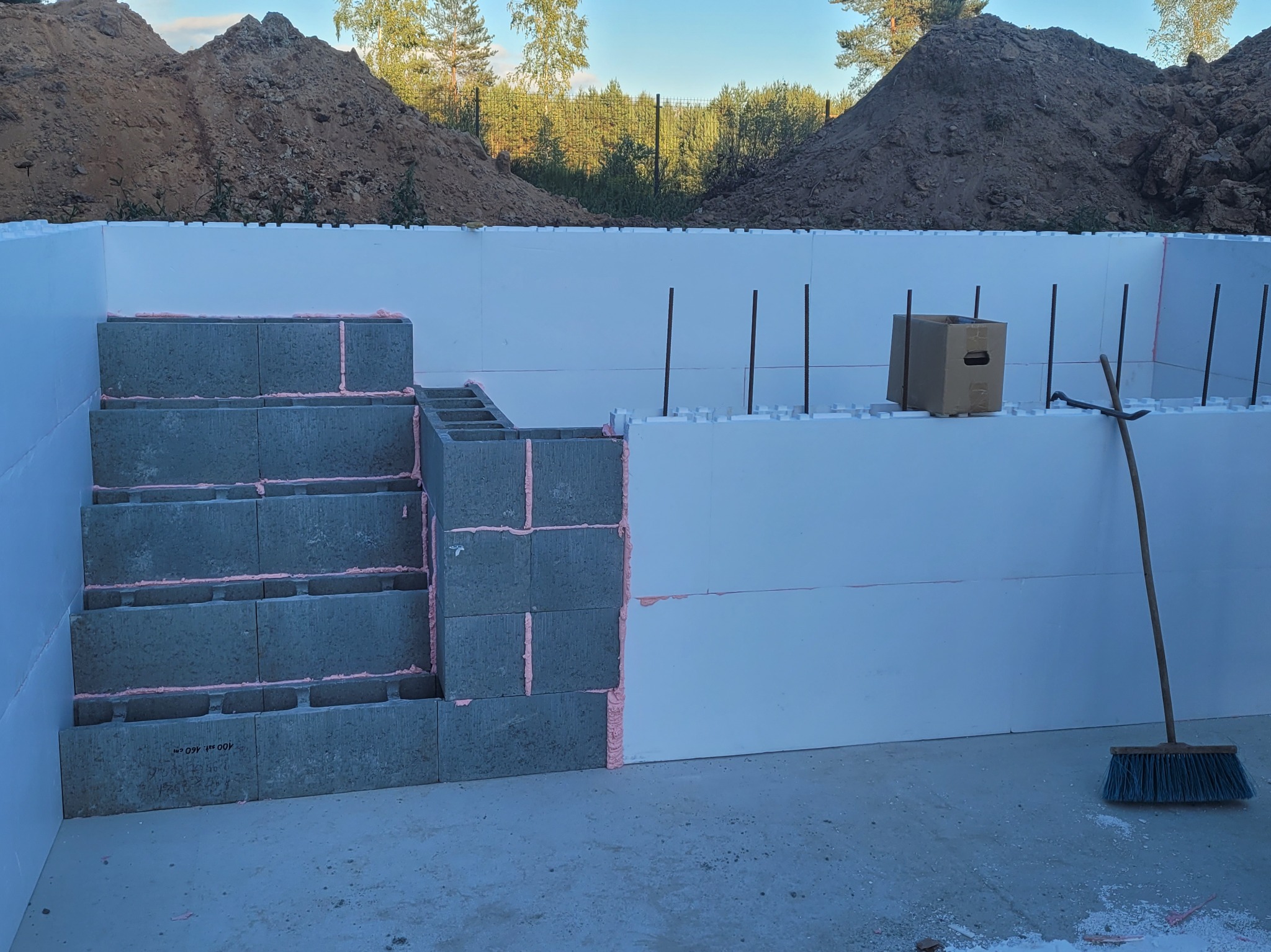 Budowa basenu: ściany z białego styropianu, betonowe schody z różową pianką montażową. W tle hałda ziemi i drzewa. Widoczna kartonowa skrzynka i miotła.