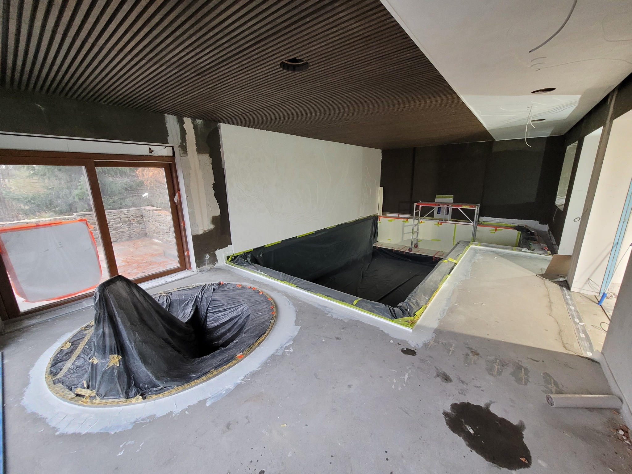 Wnętrze w trakcie budowy basenu: wykopany dół wyłożony folią, obok jacuzzi przykryte folią ochronną, widoczne narzędzia i materiały budowlane.
