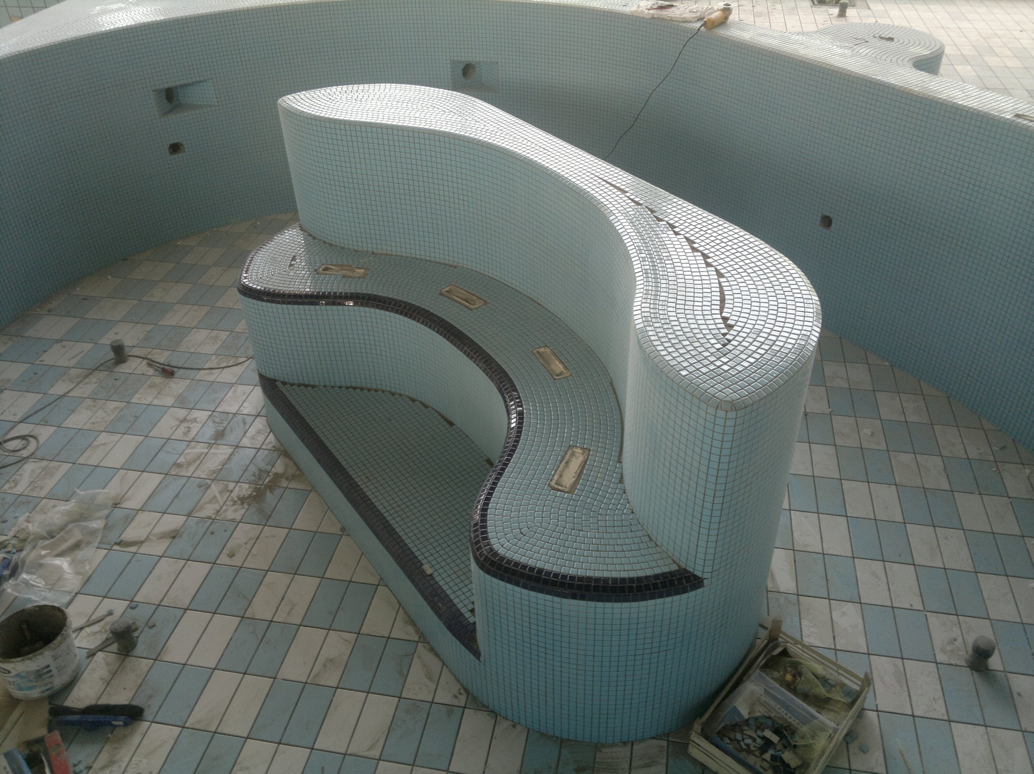 Niebieska mozaika w basenie, z widocznym siedziskiem wykończonym drobnymi płytkami. Prace wykończeniowe w toku, widoczne narzędzia i materiały budowlane.