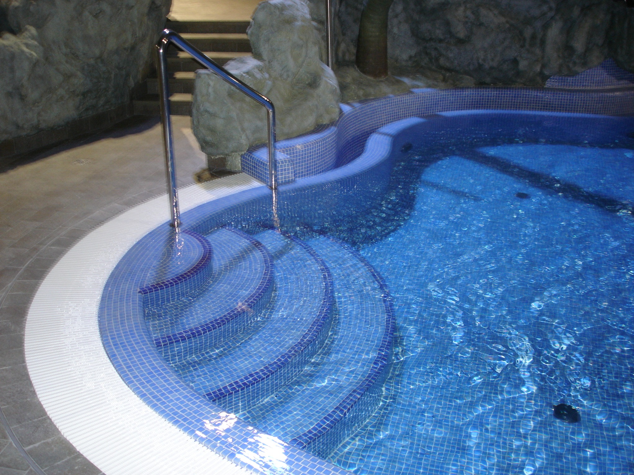 Błękitny basen z mozaiką i schodami, otoczony imitacją skał. Woda delikatnie faluje, odbijając światło. Metalowa poręcz przy wejściu do basenu.
