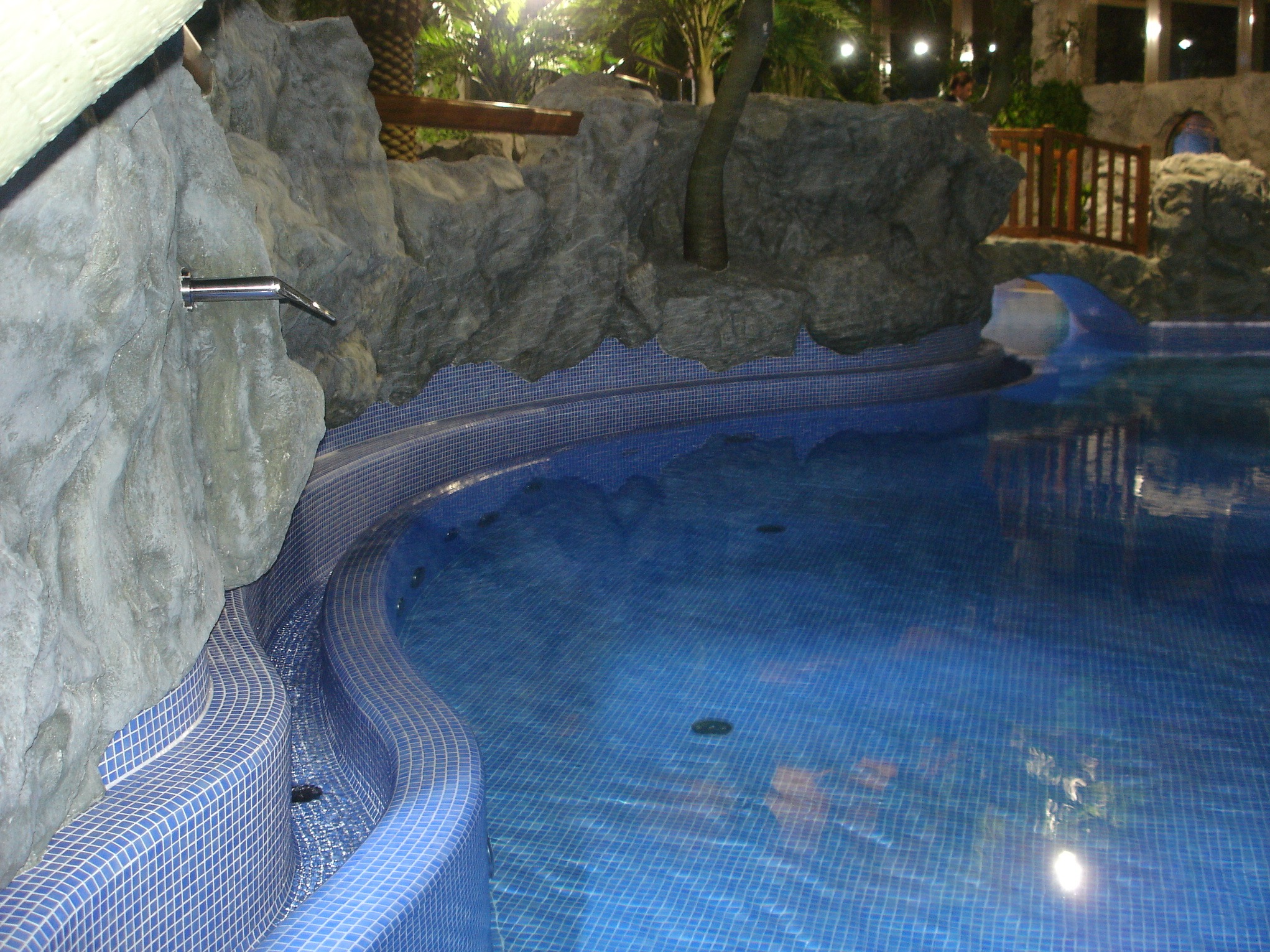 Nastrojowy basen z mozaiką i sztuczną skałą, oświetlony punktowo, z elementami drewnianej balustrady w tle. Woda w basenie ma intensywny, niebieski odcień.