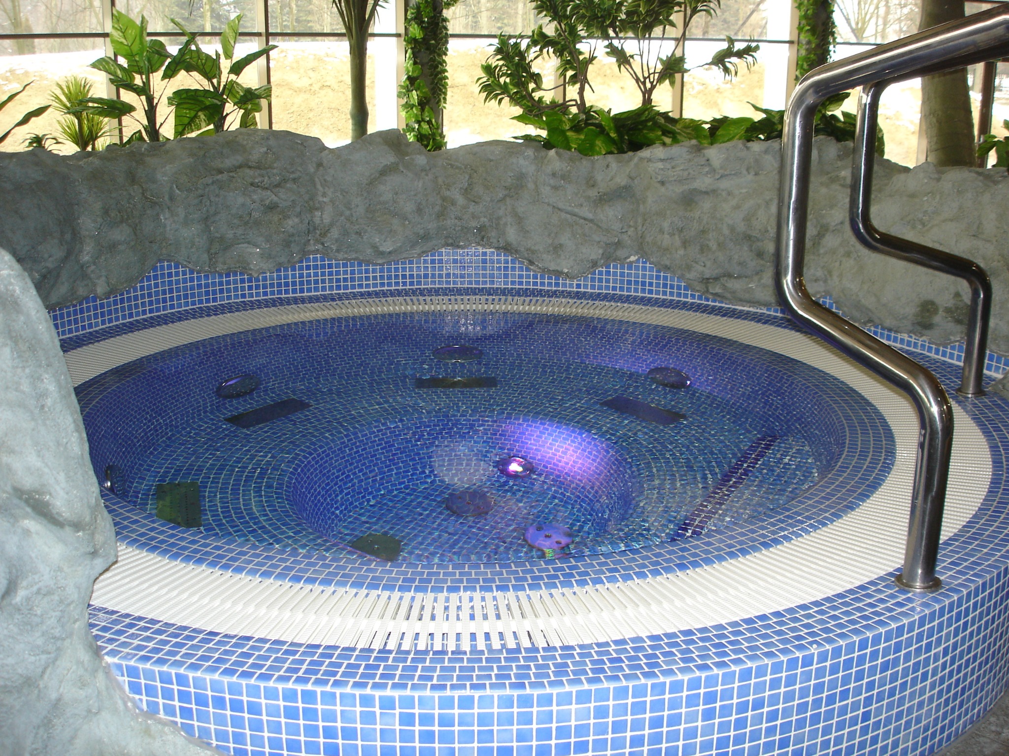 Okrągły basen z niebieskiej mozaiki, otoczony sztuczną skałą i zielenią. Metalowa poręcz ułatwia wejście. Woda w basenie delikatnie faluje, odbijając światło.