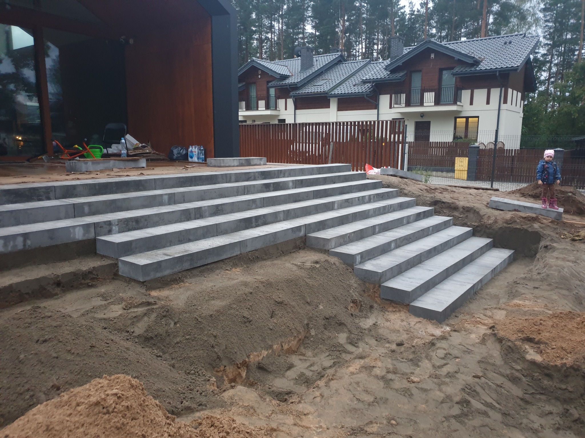 Schody z szarych, betonowych bloków prowadzące do nowoczesnego domu. W tle widoczny las i częściowo ogrodzenie. Na pierwszym planie ziemia i mała dziewczynka.