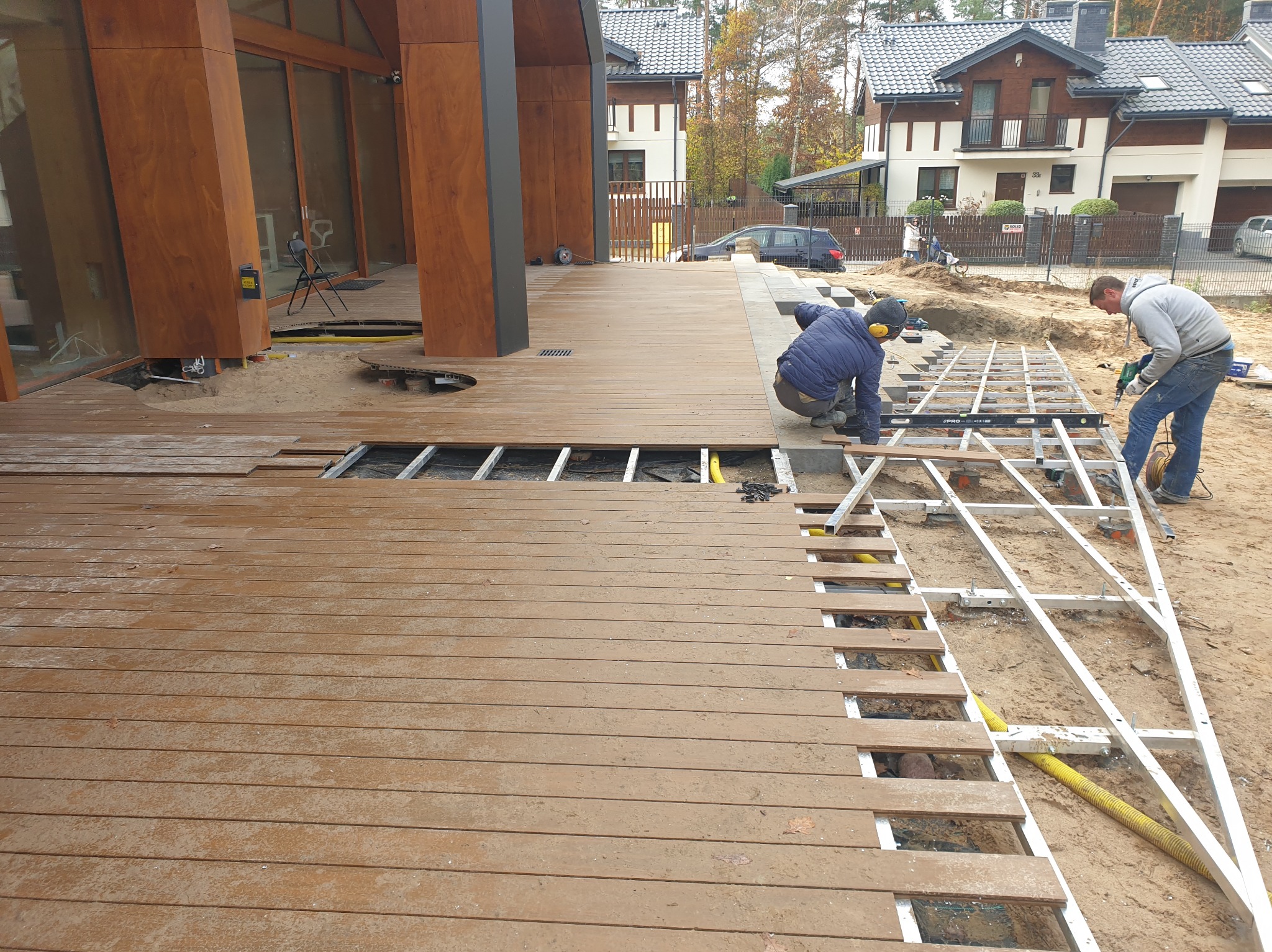 Montaż drewnianego tarasu przy domu jednorodzinnym. Widoczne deski tarasowe, konstrukcja nośna i pracownicy podczas układania. Częściowo ułożona podłoga tarasu.
