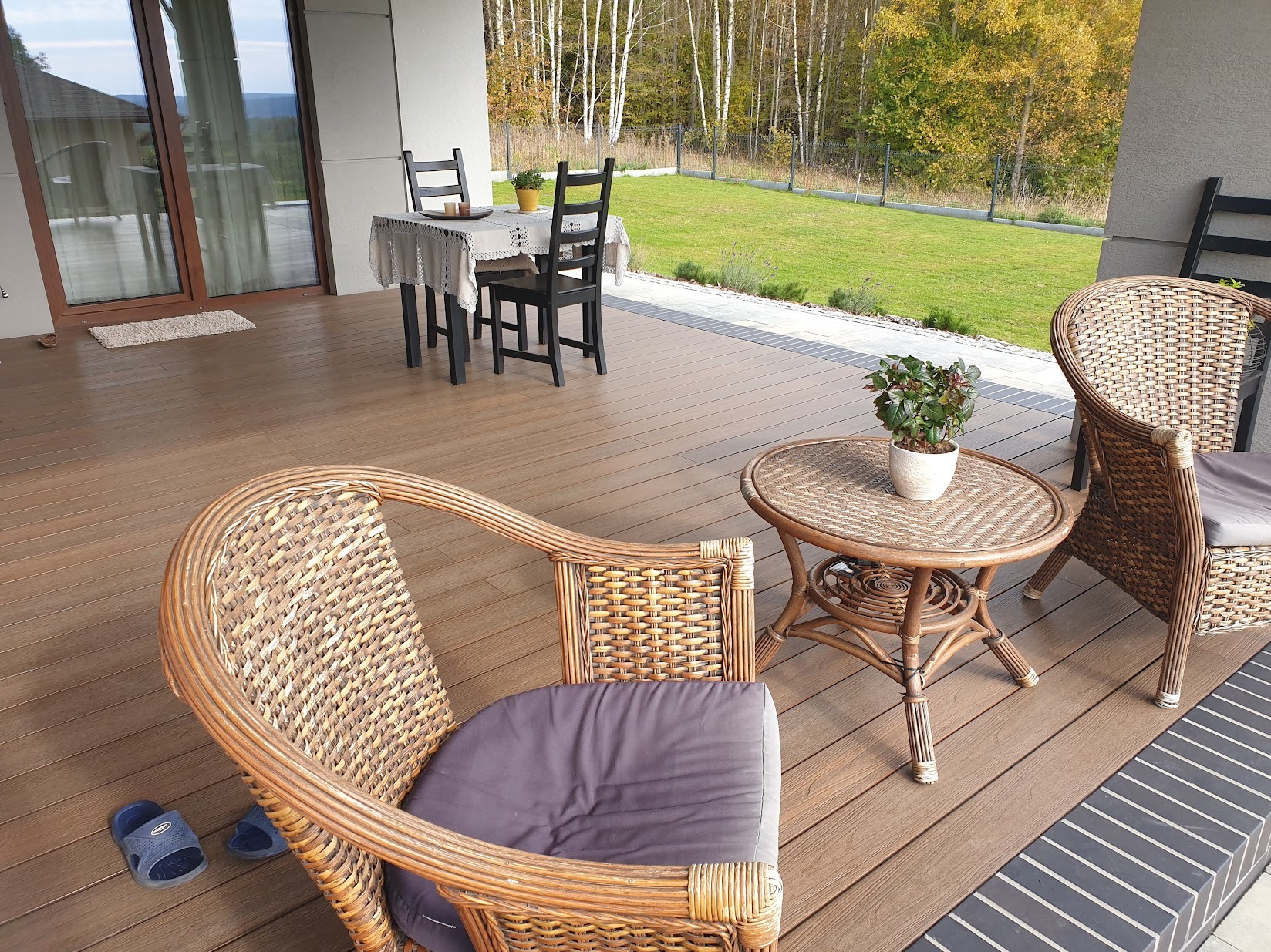 Drewniany taras z rattanowymi meblami ogrodowymi i widokiem na zieleń. Stół z krzesłami w tle. Ujęcie z niskiej perspektywy, podkreślające detale wykończenia tarasu.