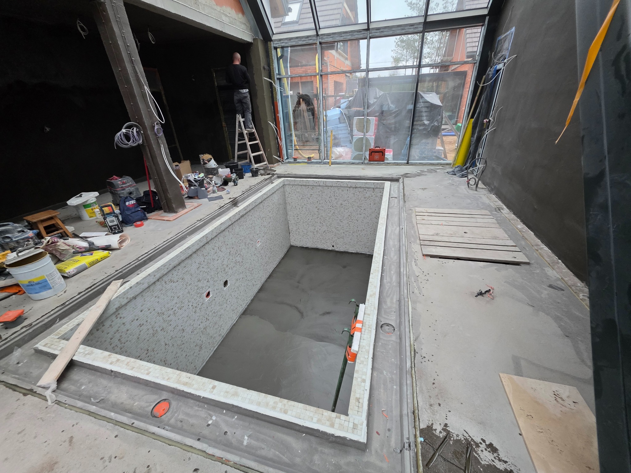 Wnętrze basenu w trakcie budowy z mozaiką szkliwioną, widoczne narzędzia i pracownik na drabinie. Duże okno z widokiem na zewnątrz. Etap doszczelniania przepustów w żelbecie.