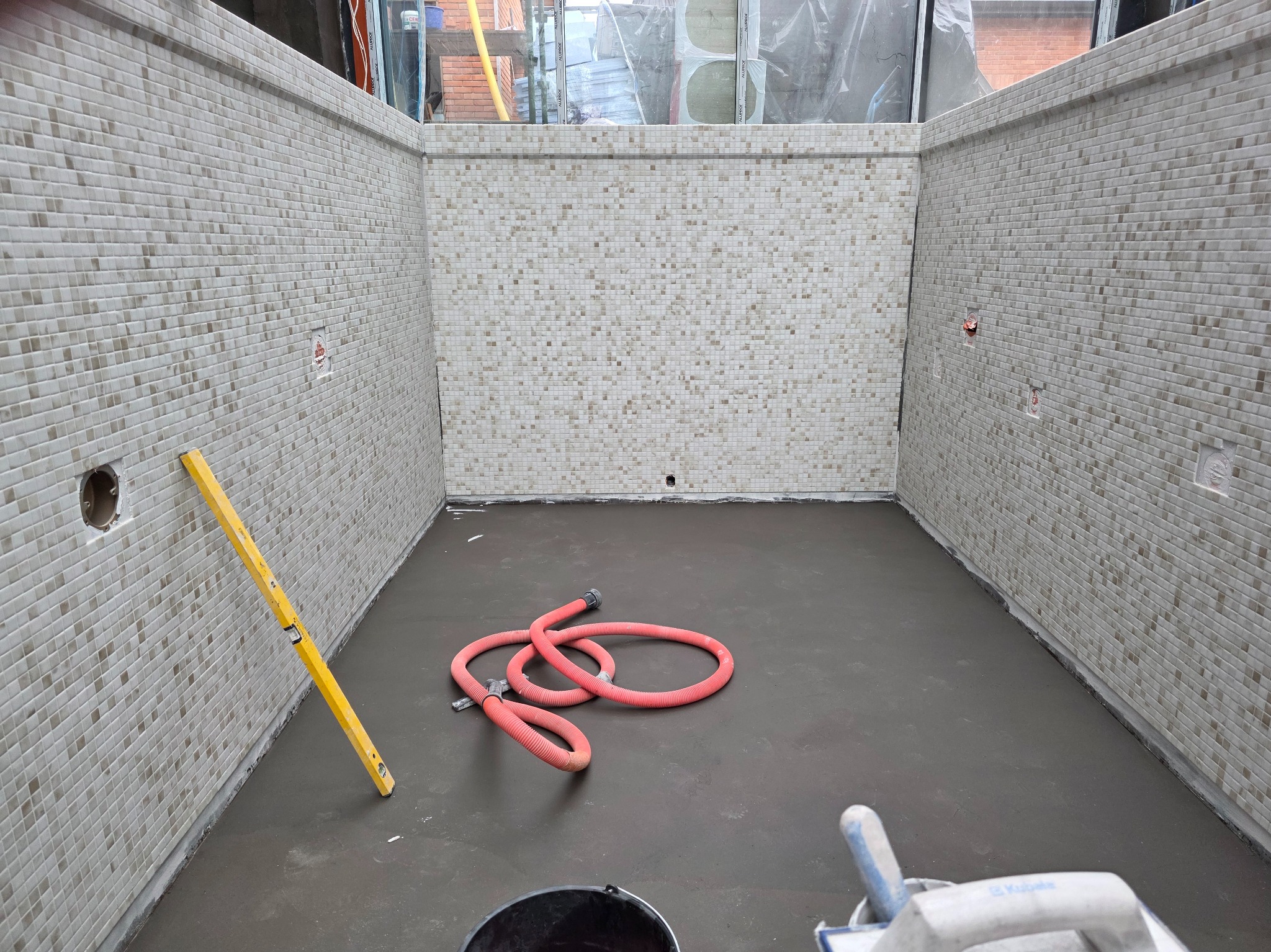 Wnętrze basenu w trakcie budowy: wyłożone mozaiką ściany, wylewka na dnie, poziomica i wąż. Widoczne przygotowania do dalszych prac wykończeniowych.