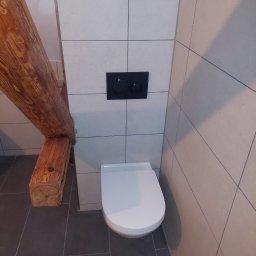 Majster Lenar - Nowoczesna toaleta podwieszana z czarnym panelem spłukującym na tle jasnych płytek, z widoczną drewnianą belką stropową. Minimalistyczny design w łazience.