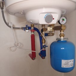 Majster Lenar - Biały bojler Galmet z niebieskim zbiornikiem wyrównawczym i instalacją hydrauliczną w pomieszczeniu. Widoczne rury w izolacji i manometr.