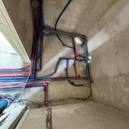 Majster Lenar - Rozbudowana instalacja wodna i grzewcza z rur PEX w kolorach niebieskim i czerwonym, zamocowana do surowej ściany w trakcie prac wykończeniowych.