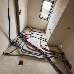 Majster Lenar - Surowy stan łazienki z oknem dachowym. Widoczne rury instalacji wodnej (czerwone i niebieskie) oraz kanalizacyjne, ułożone na podłodze przed wylaniem posadzki.