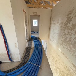 Majster Lenar - Niebieskie i szare rury instalacyjne ułożone wzdłuż korytarza w surowym stanie, widok z góry, drewniana konstrukcja dachu w tle, ściany w trakcie wykończenia.