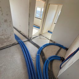 Majster Lenar - Niebieskie rury instalacyjne na podłodze w surowym wnętrzu budynku we Wrocławiu. Widoczne kable i przygotowane otwory drzwiowe. Instalacja przed wylewką.