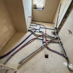 Majster Lenar - Rozłożone rury instalacji wodnej i kanalizacyjnej w surowym pomieszczeniu z oknem. Widoczne rury czerwone, niebieskie i szare. Częściowo widoczne buty w dolnej części kadru.