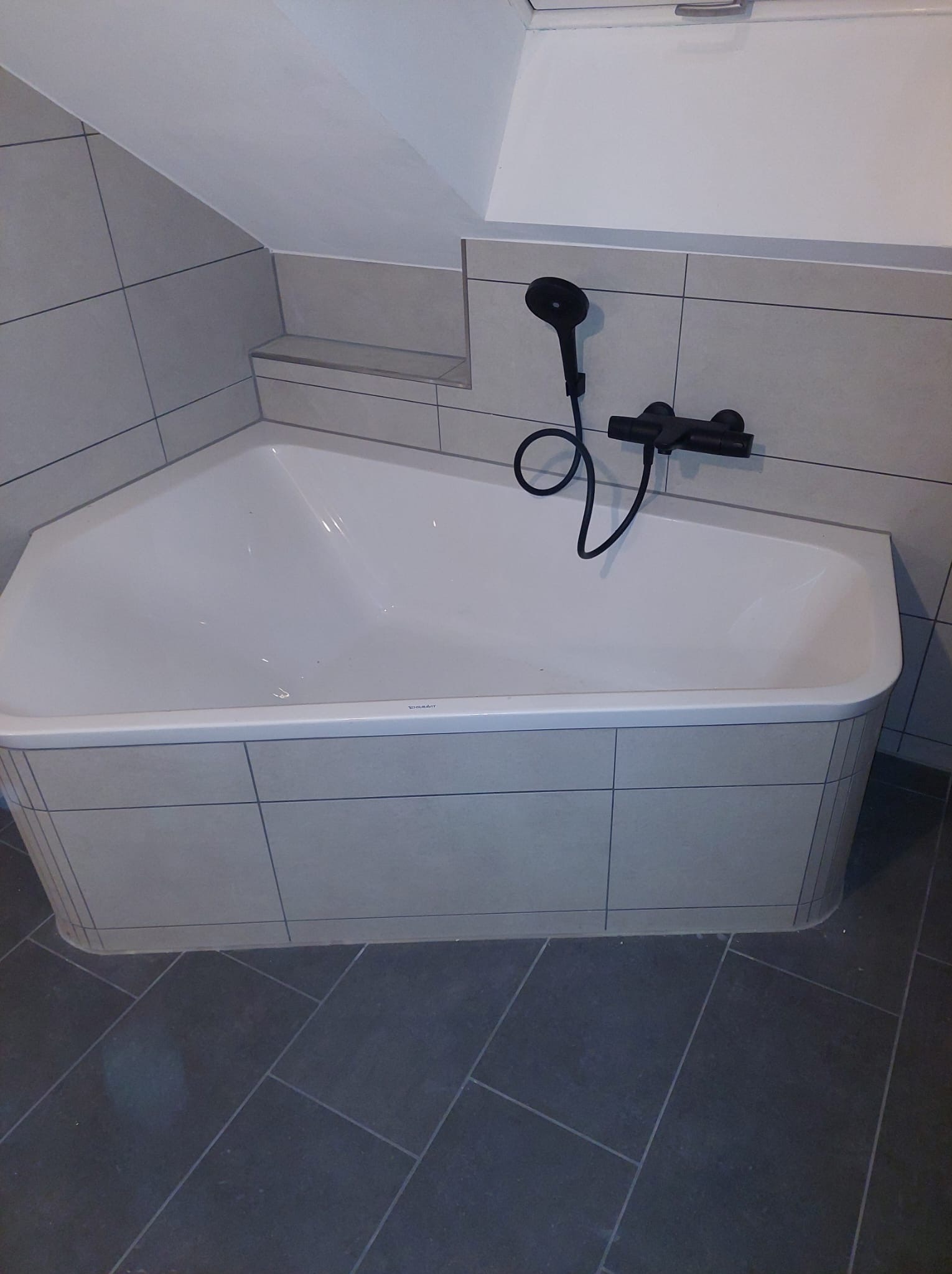 Narożna wanna z czarną armaturą i szarymi płytkami w łazience na poddaszu. Ukończona instalacja sanitarna z widocznym odpływem.