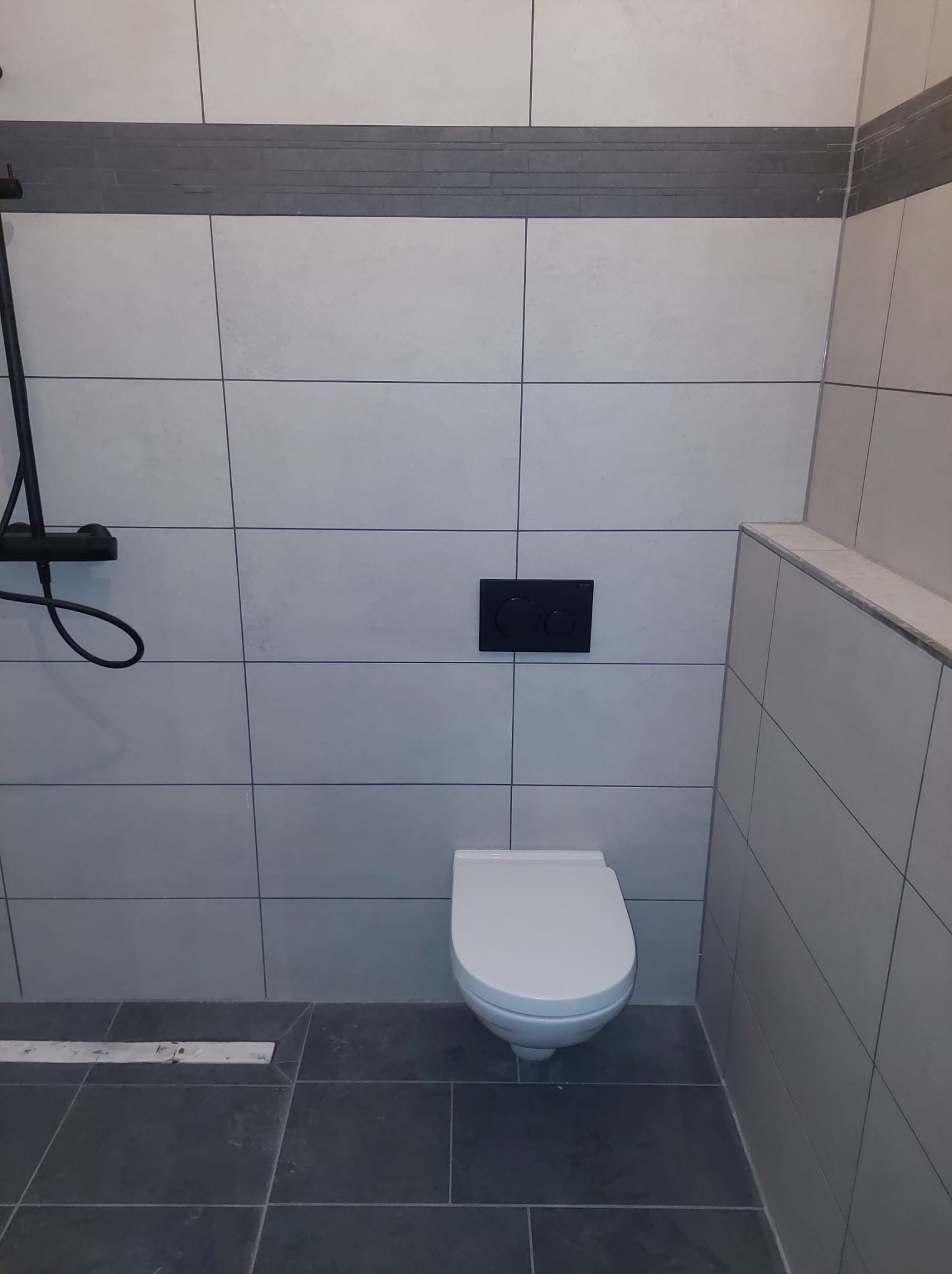 Nowoczesna toaleta z białym kompaktem WC i czarnym przyciskiem spłukującym na tle jasnych, kwadratowych płytek. Widoczny fragment czarnej armatury prysznicowej.