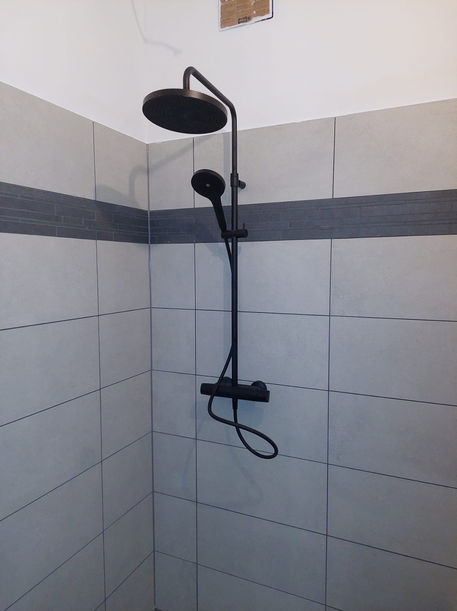 Nowoczesny zestaw prysznicowy w kolorze czarnym, zamontowany na ścianie wyłożonej szarymi płytkami z dekoracyjnym pasem. Minimalistyczny design, widok z dołu.