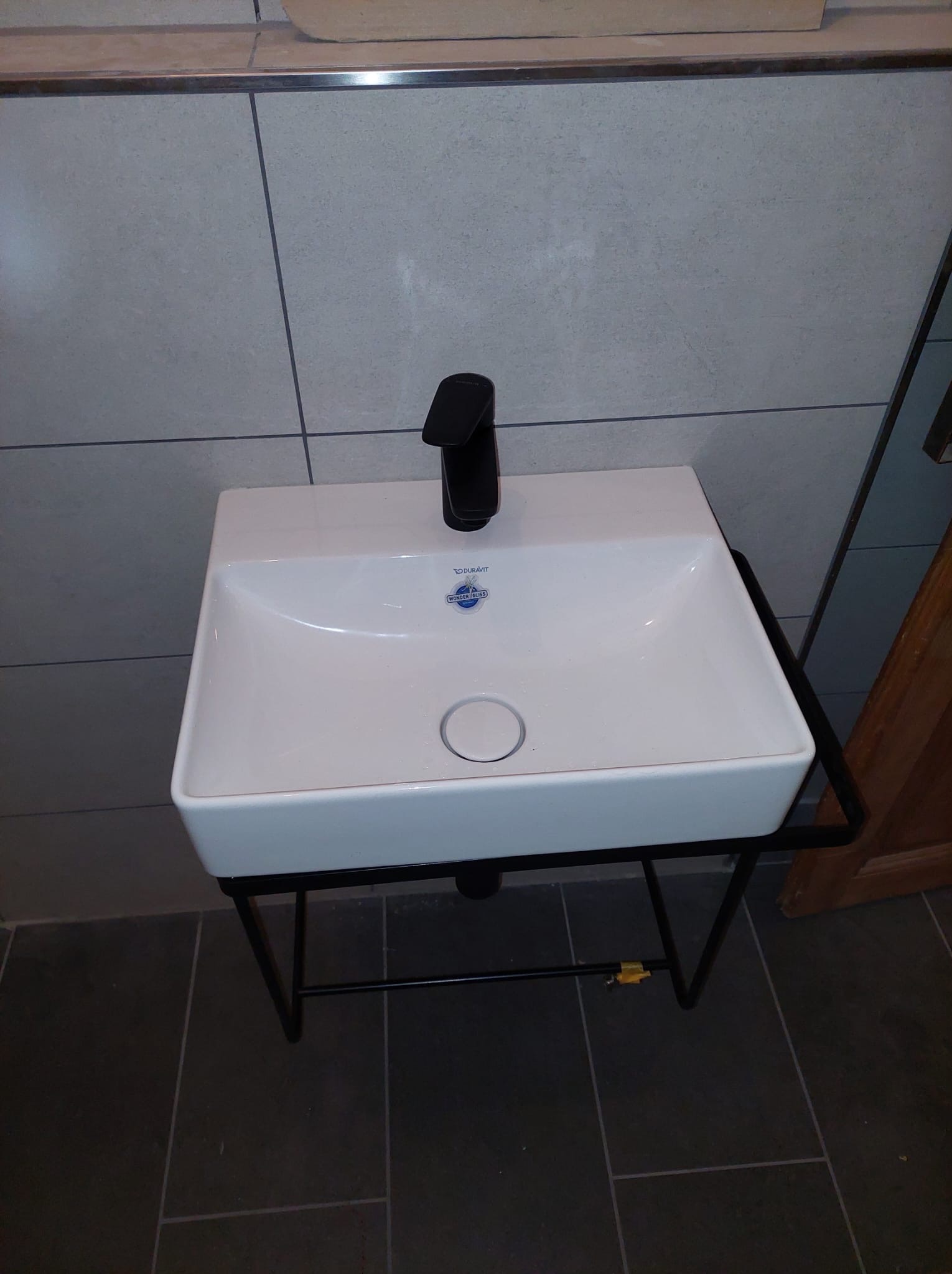 Nowoczesna, biała umywalka Duravit z czarną baterią, zamontowana na metalowym stelażu w łazience z szarymi płytkami. Minimalistyczny design.