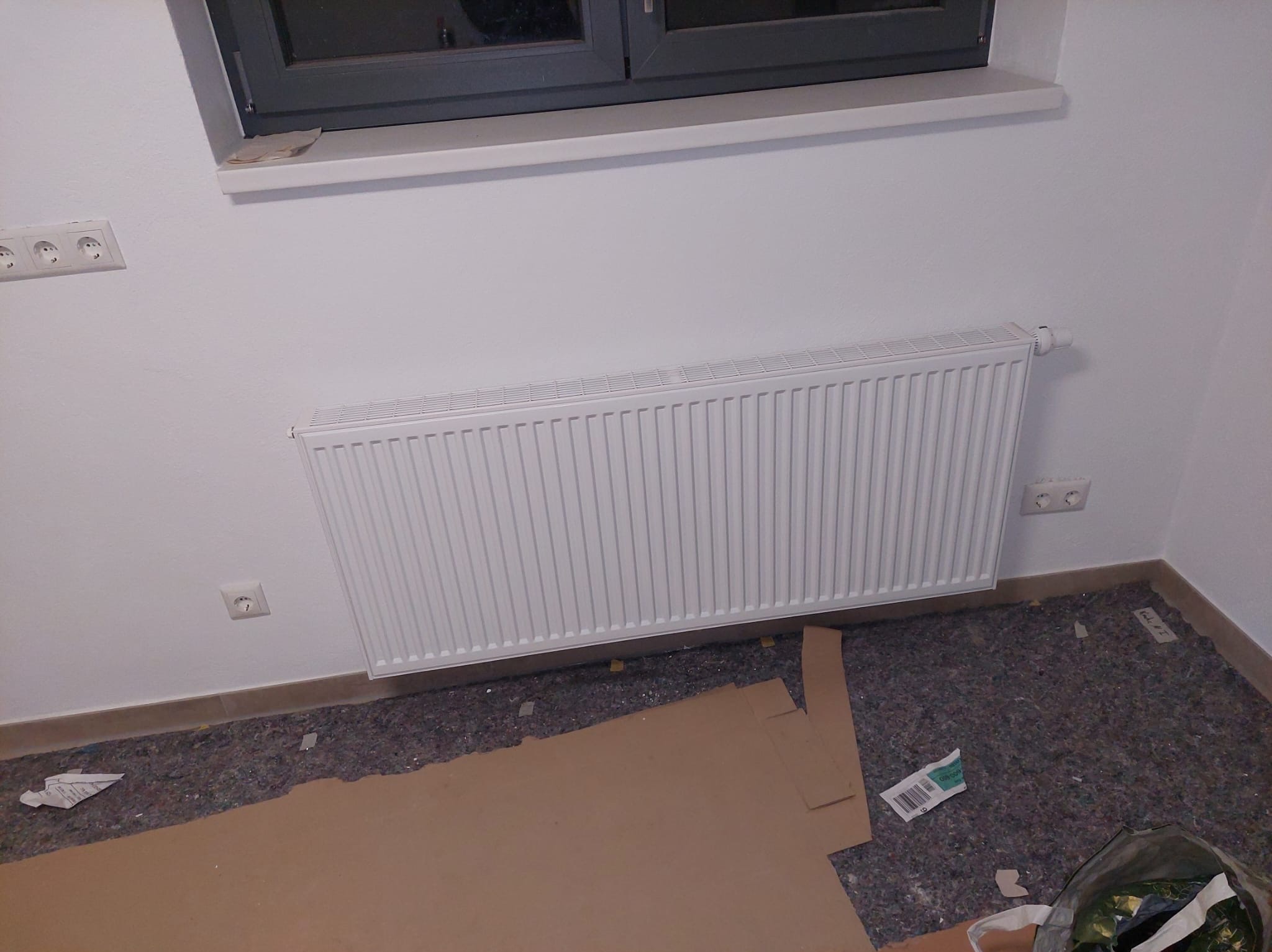 Biały, nowy grzejnik panelowy zamontowany pod oknem na białej ścianie. Na podłodze widoczne materiały zabezpieczające podłoże podczas prac instalacyjnych.