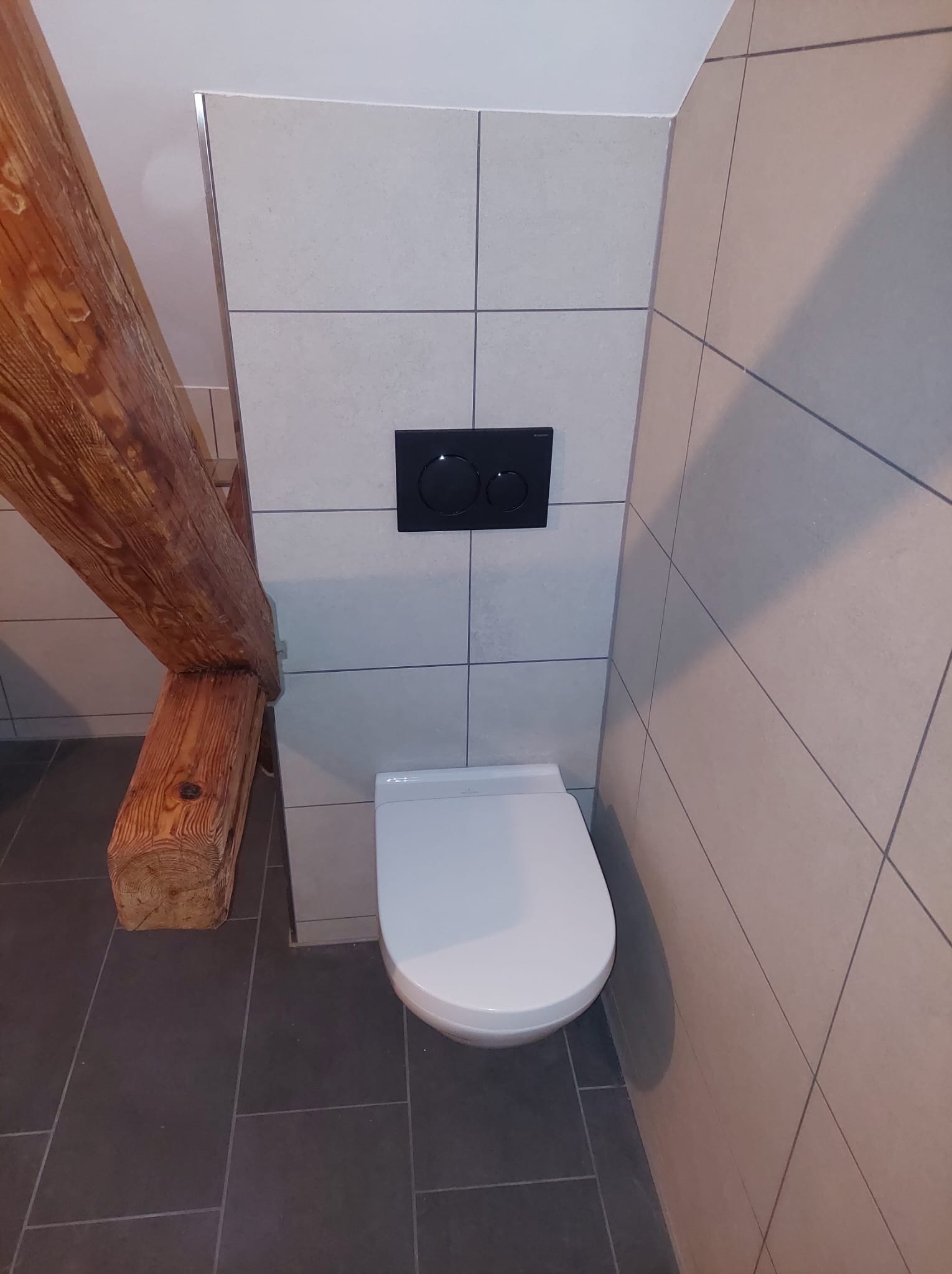 Nowoczesna toaleta podwieszana z czarnym panelem spłukującym na tle jasnych płytek, z widoczną drewnianą belką stropową. Minimalistyczny design w łazience.