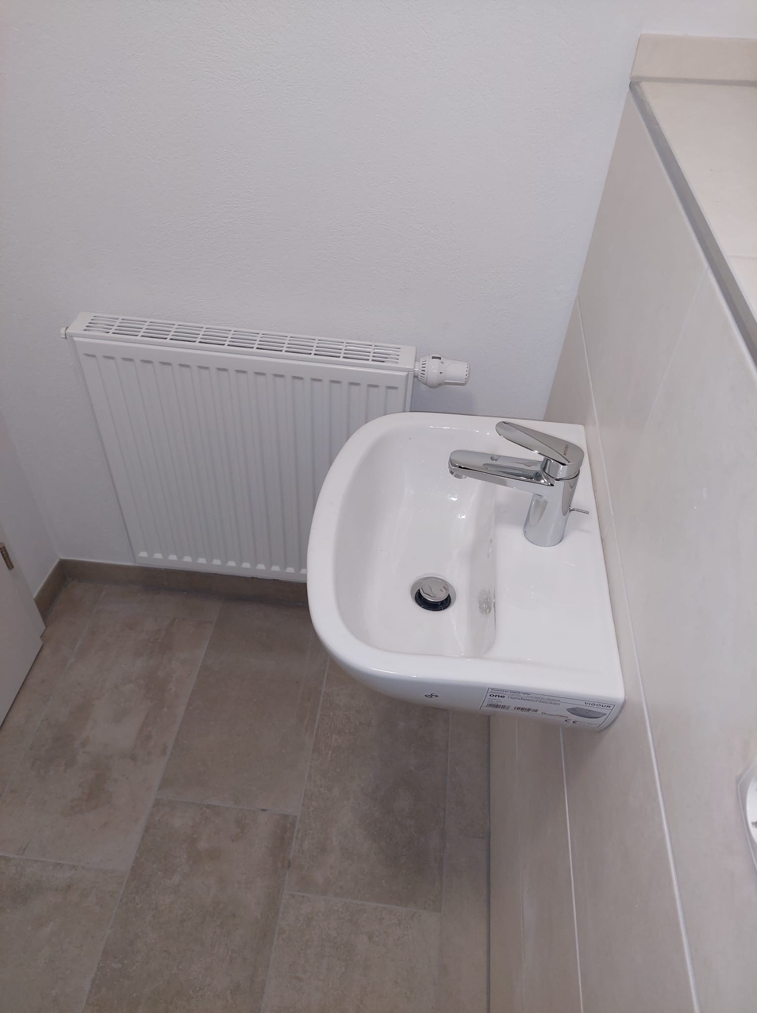 Biała umywalka z chromowaną baterią w łazience z grzejnikiem. Widoczne płytki na ścianie i podłodze. Ujęcie z góry, prezentujące detale instalacji.