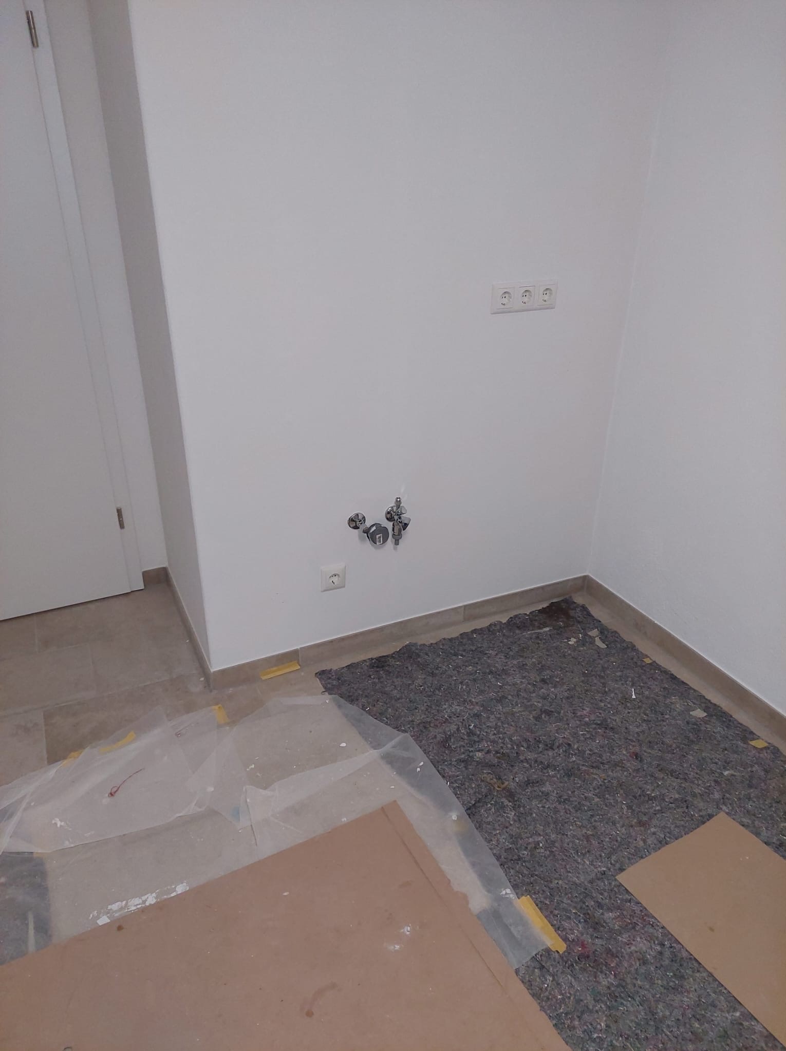 Przygotowanie do prac hydraulicznych: widoczne przyłącze wodne na ścianie, zabezpieczona folią podłoga i płyty kartonowe. Gniazdka elektryczne nad instalacją.