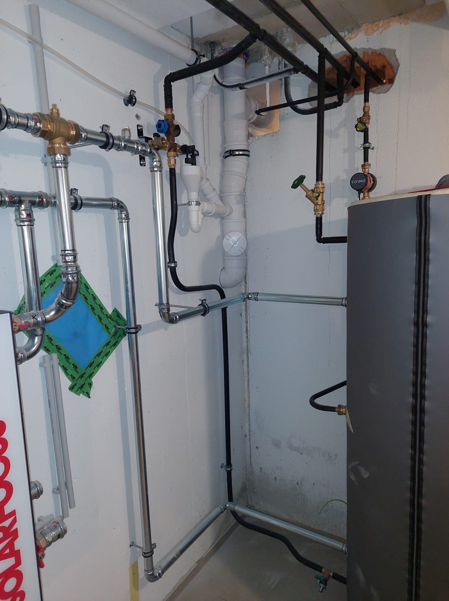 Złożona instalacja grzewcza z rurami miedzianymi i czarnymi, zaworami oraz pompą obiegową. Widoczne elementy mocujące i izolacja. Ściana z surowym wykończeniem.