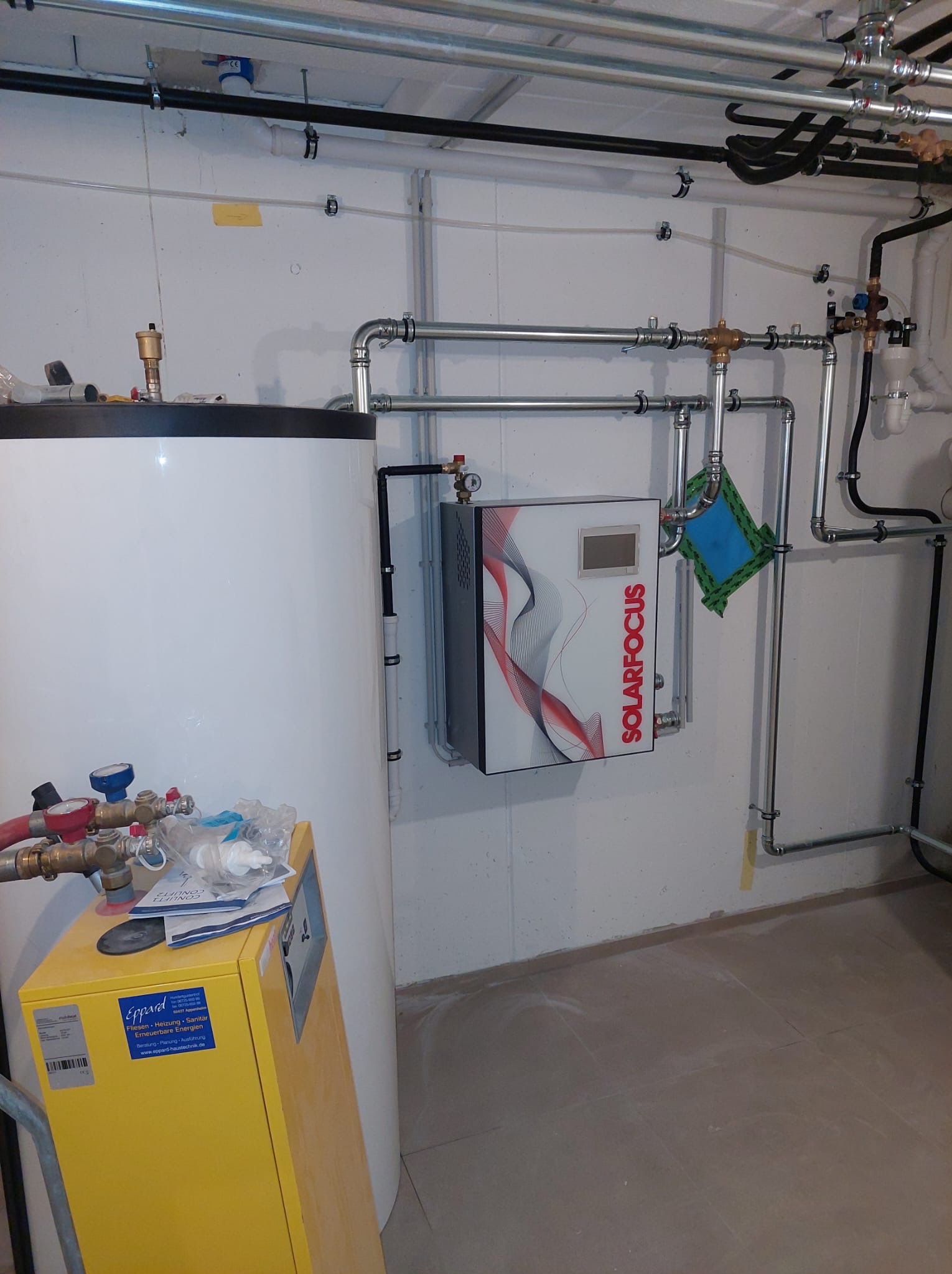 Biały zasobnik ciepłej wody z żółtym piecem Eppard i panelem sterowania Solarfocus na ścianie. Widoczne rury instalacji grzewczej.