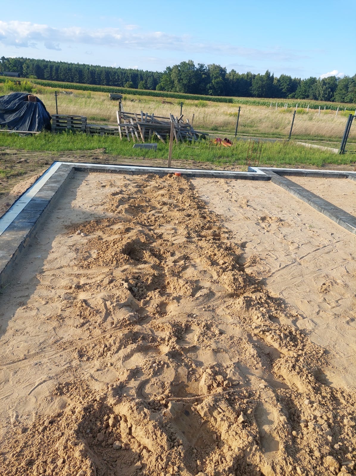 Przygotowanie terenu pod budowę: wyrównany piasek w obramowaniu z kostki, widoczne ślady prac ziemnych, w tle łąka i las. Dzień słoneczny.