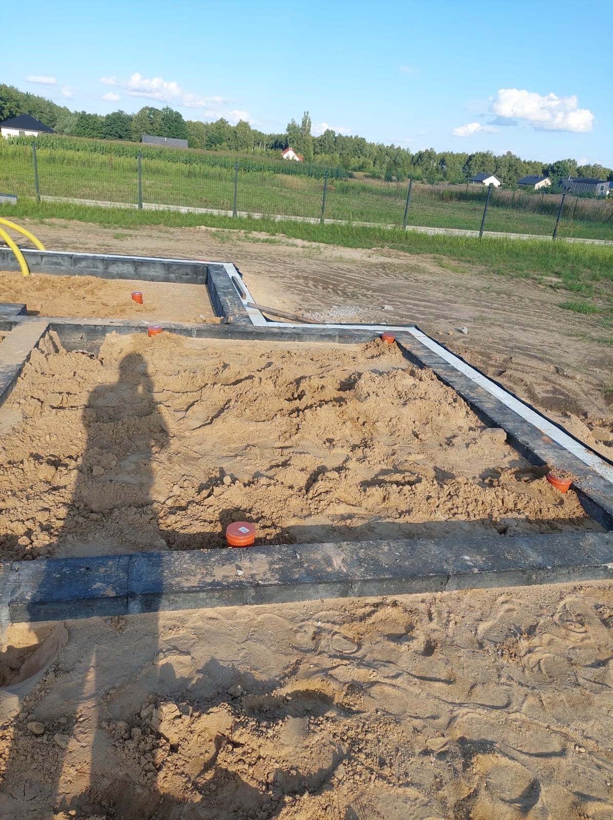 Fundamenty domu w trakcie budowy: piasek wewnątrz obramowania z kostki, wystające rury. W tle trawa i drzewa, cień osoby robiącej zdjęcie.