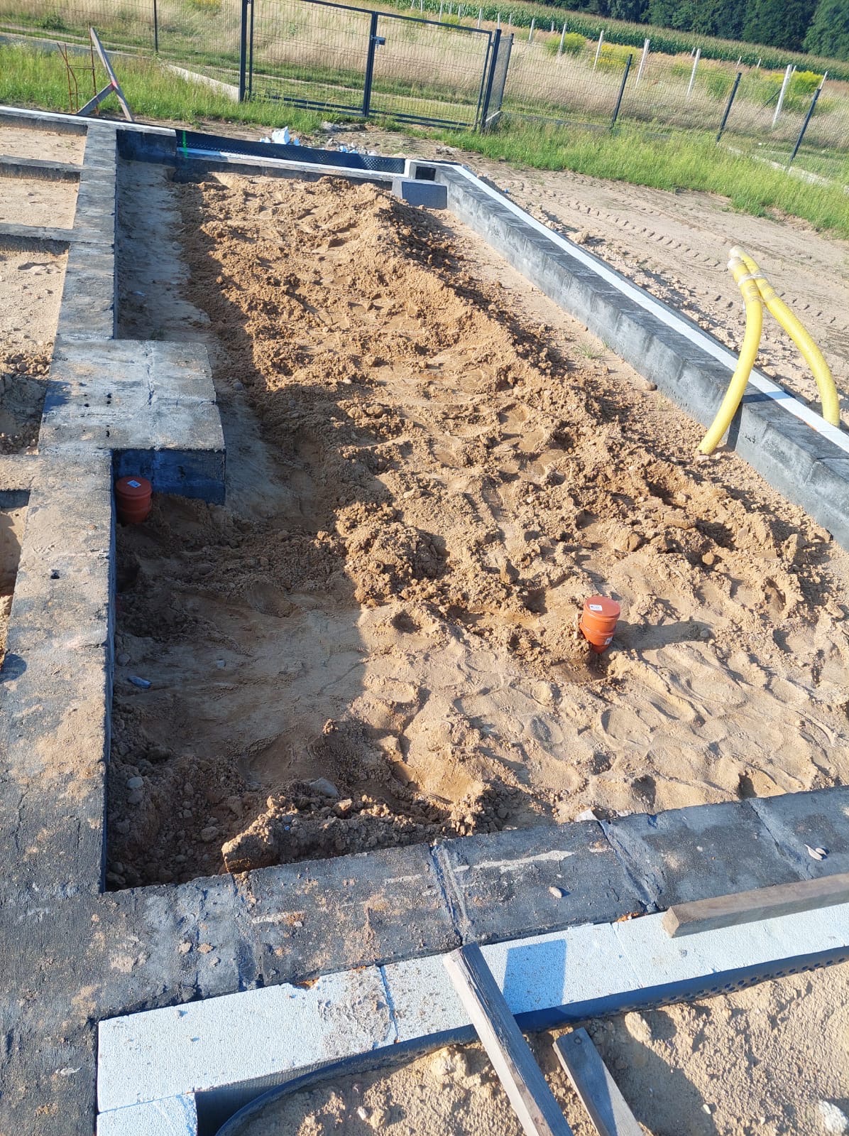 Fundamenty domu jednorodzinnego w trakcie budowy, widoczne rury instalacyjne i piasek. Ujęcie z góry, w tle ogrodzenie i zieleń.