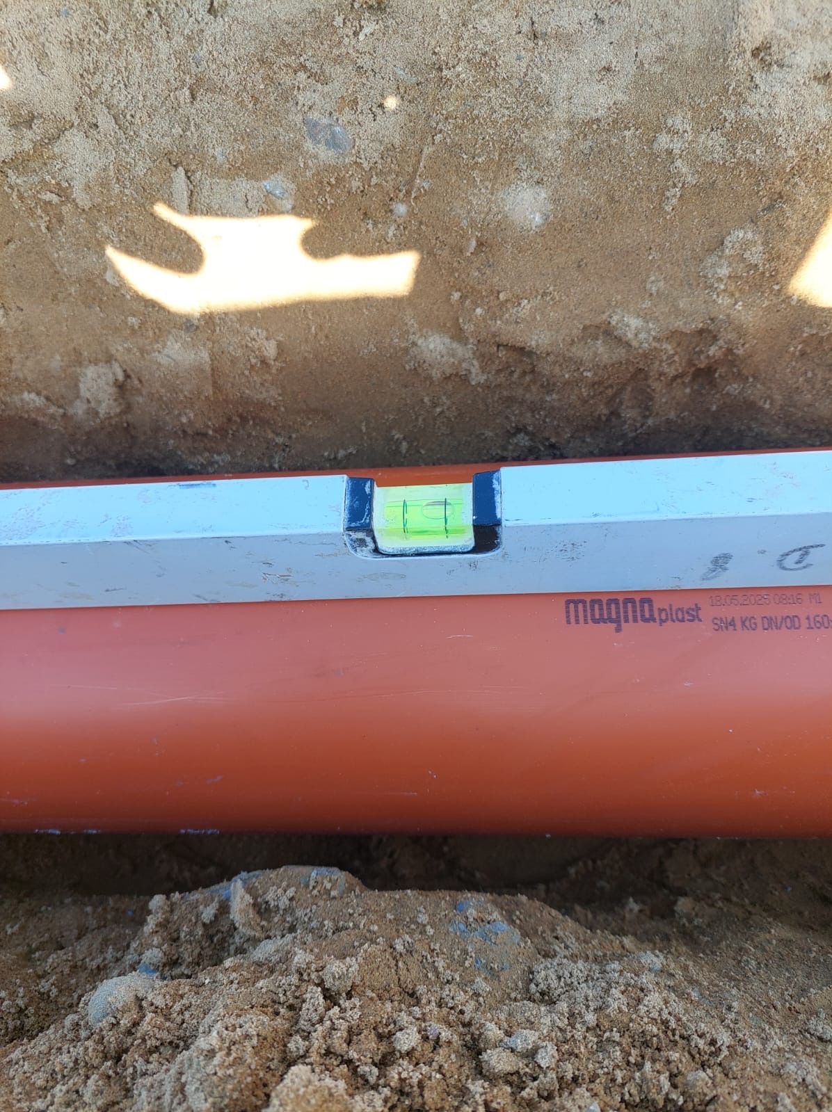 Poziomnica na pomarańczowej rurze Magna Plast w wykopie z piaskiem. Widoczny fragment rury z oznaczeniami producenta i datą. Kontrola poziomu instalacji.