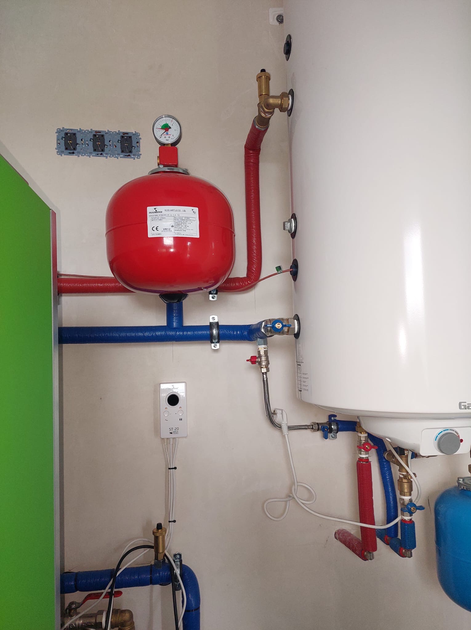 Złożona instalacja grzewcza z czerwonym zbiornikiem wyrównawczym i białym bojlerem, rury w izolacji niebieskiej i czerwonej, regulator temperatury na ścianie.